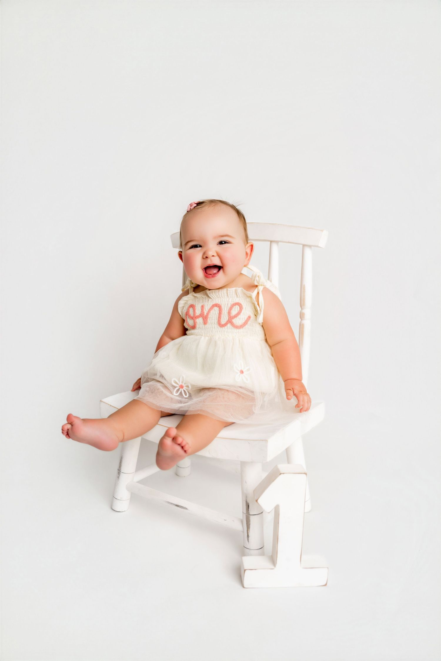 Smiling baby girl in white tutu dress with “one” bib sits on white chair celebrating her first birthday.