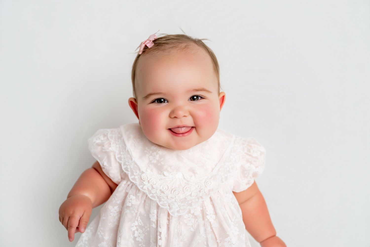 Smiling baby girl in white lace dress with pink bow headband posed against white background.