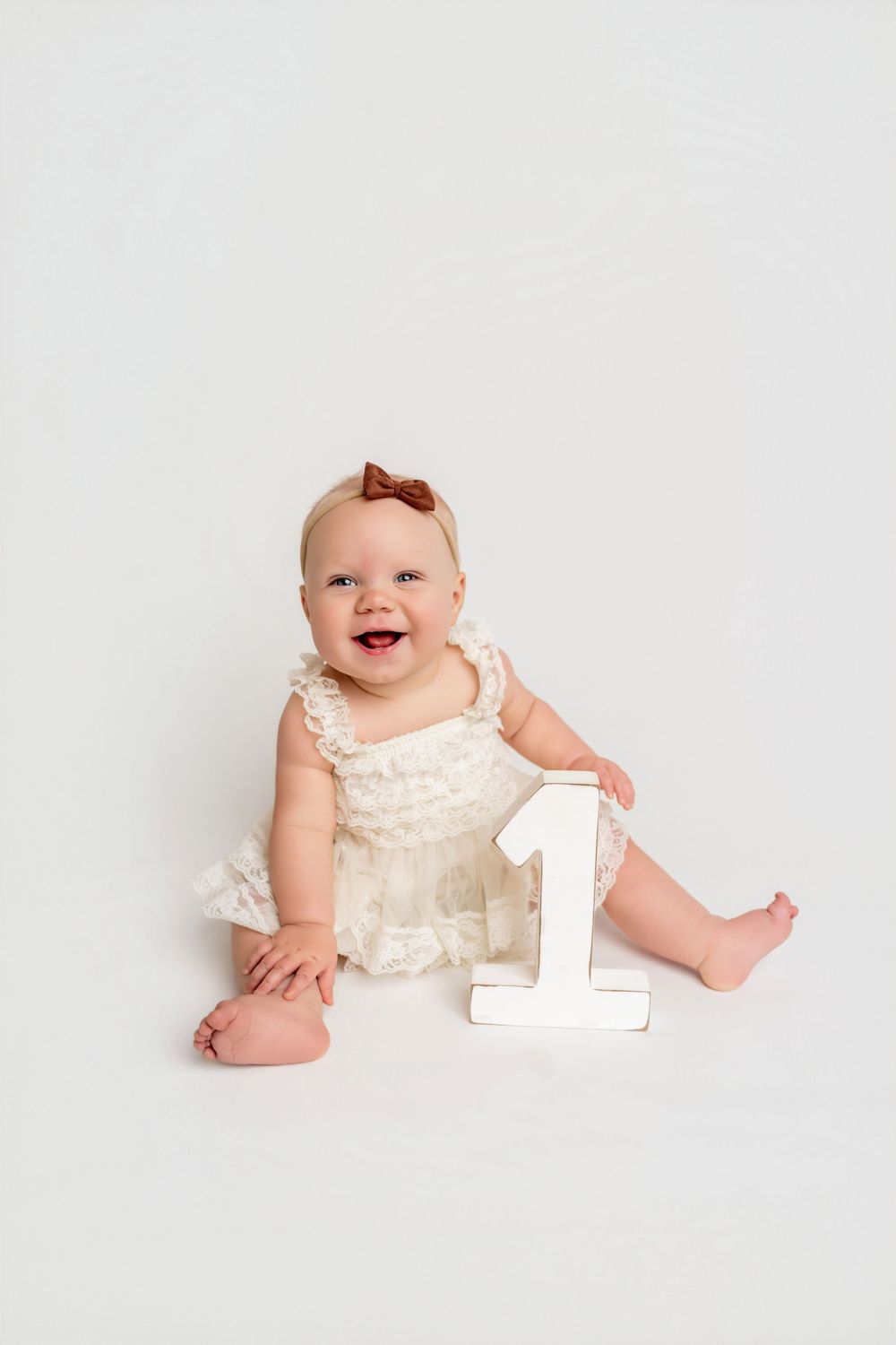 Smiling baby girl in white lace dress sitting with number 1 sign, celebrating her first birthday.