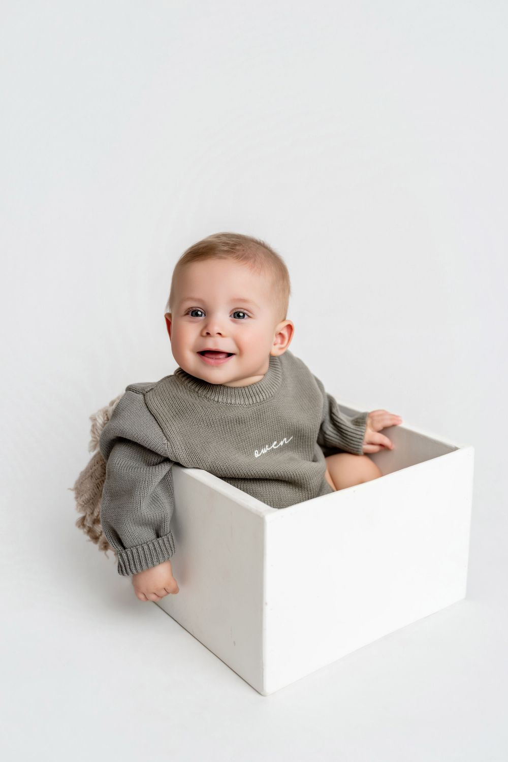Smiling baby in a sage green knit sweater sitting inside a white box against a white background.