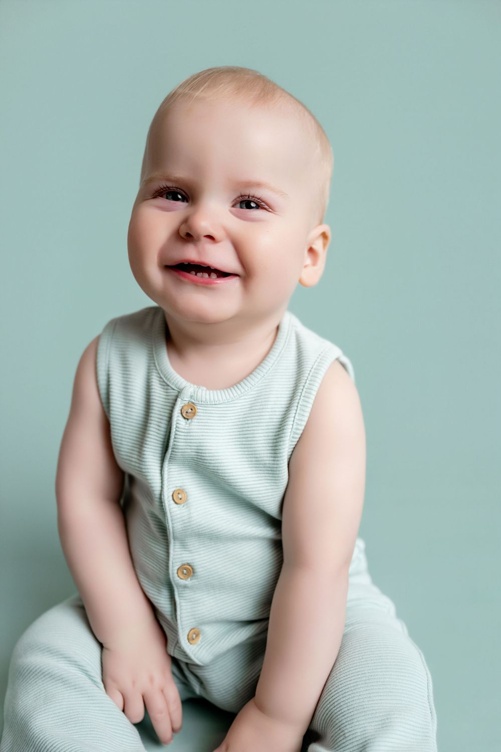 Smiling baby in a mint green sleeveless romper sitting against a light blue background.