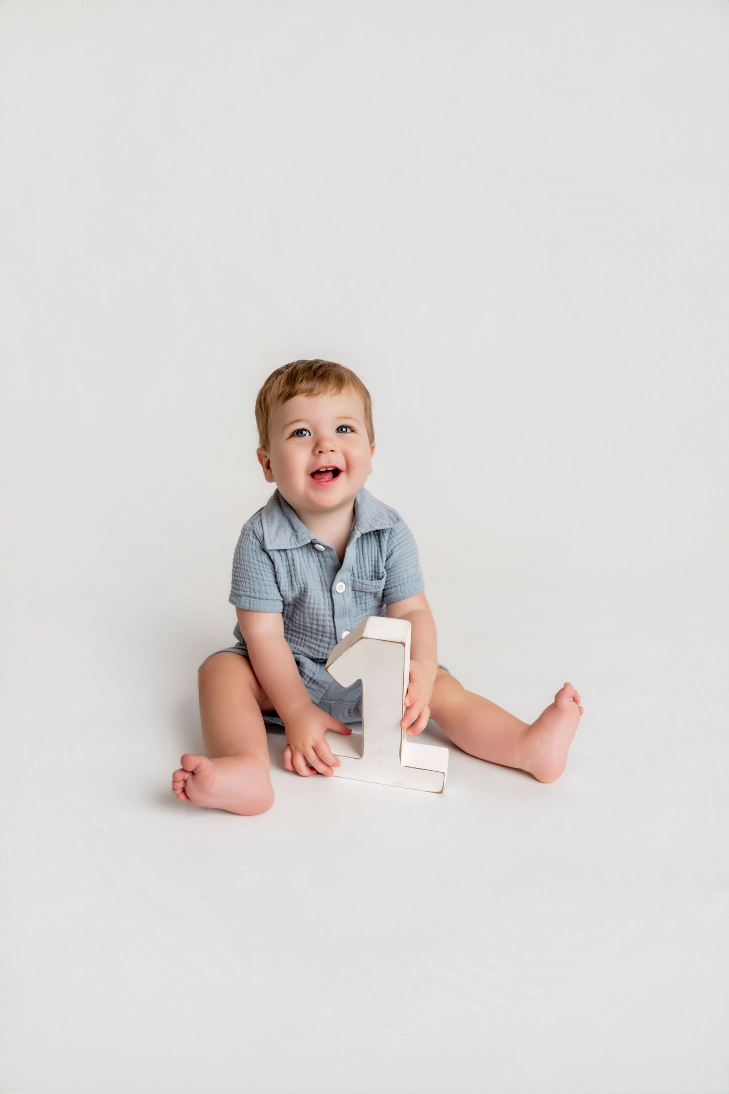 Smiling baby boy in blue romper sitting on white floor holding a wooden number one for first birthday photos.