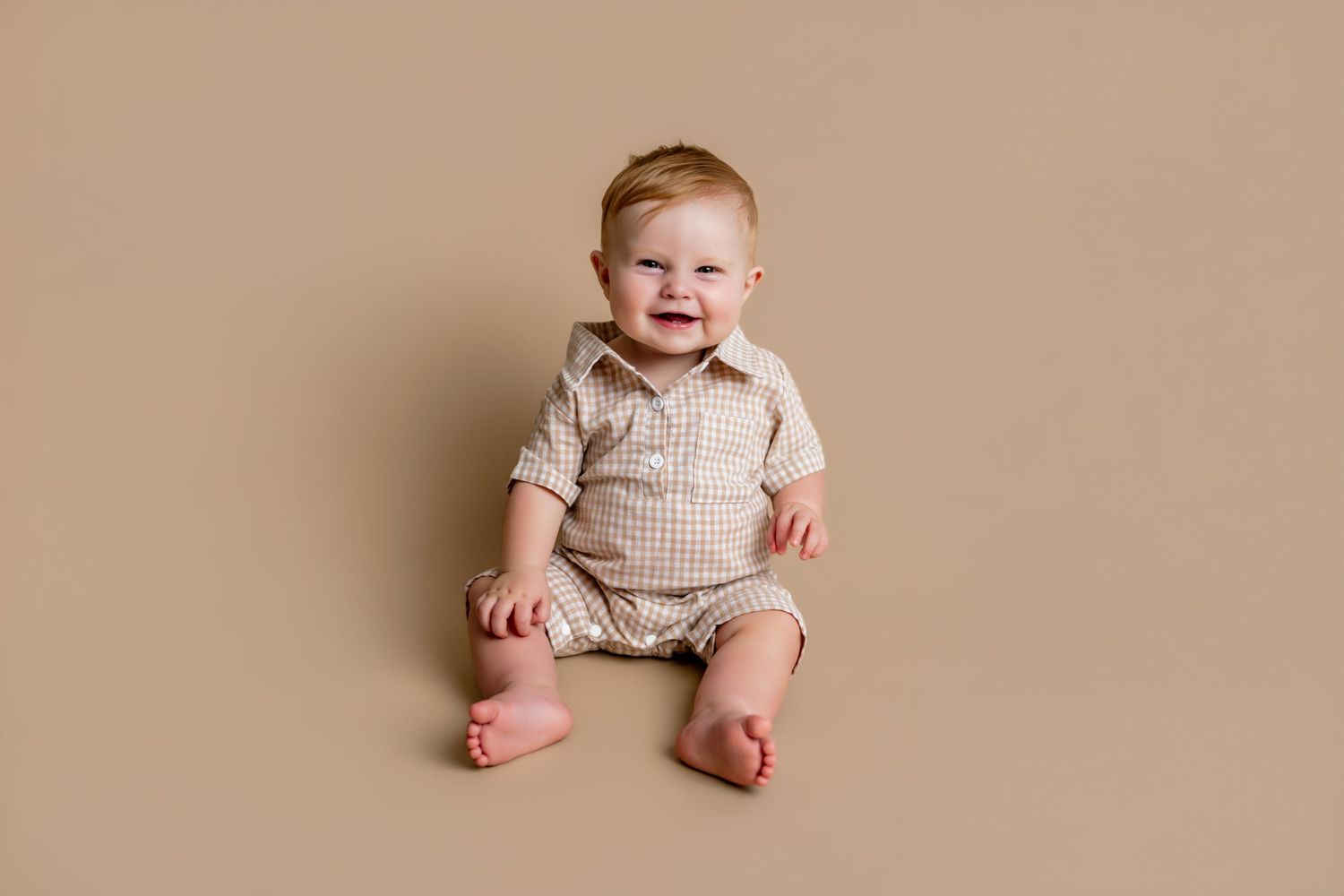 Smiling baby boy sitting on beige backdrop wearing a tan checkered romper during a milestone photo session.