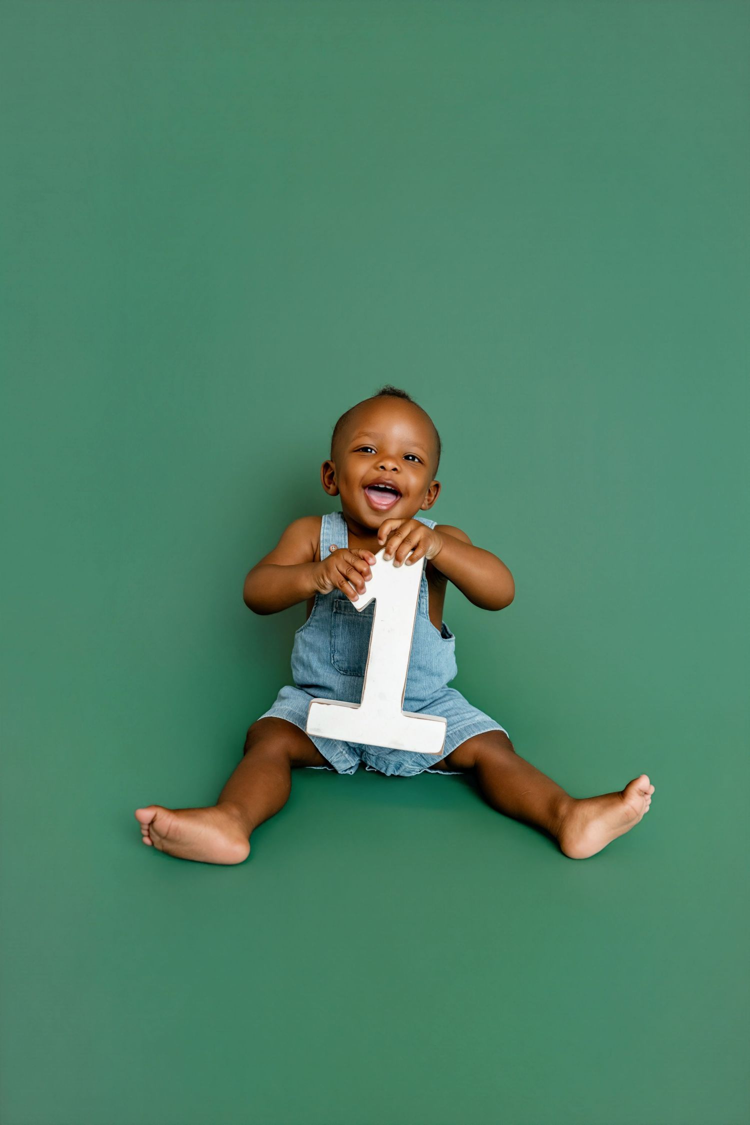 Happy Black baby in denim overalls sitting on green background holding white number 1, celebrating first birthday.