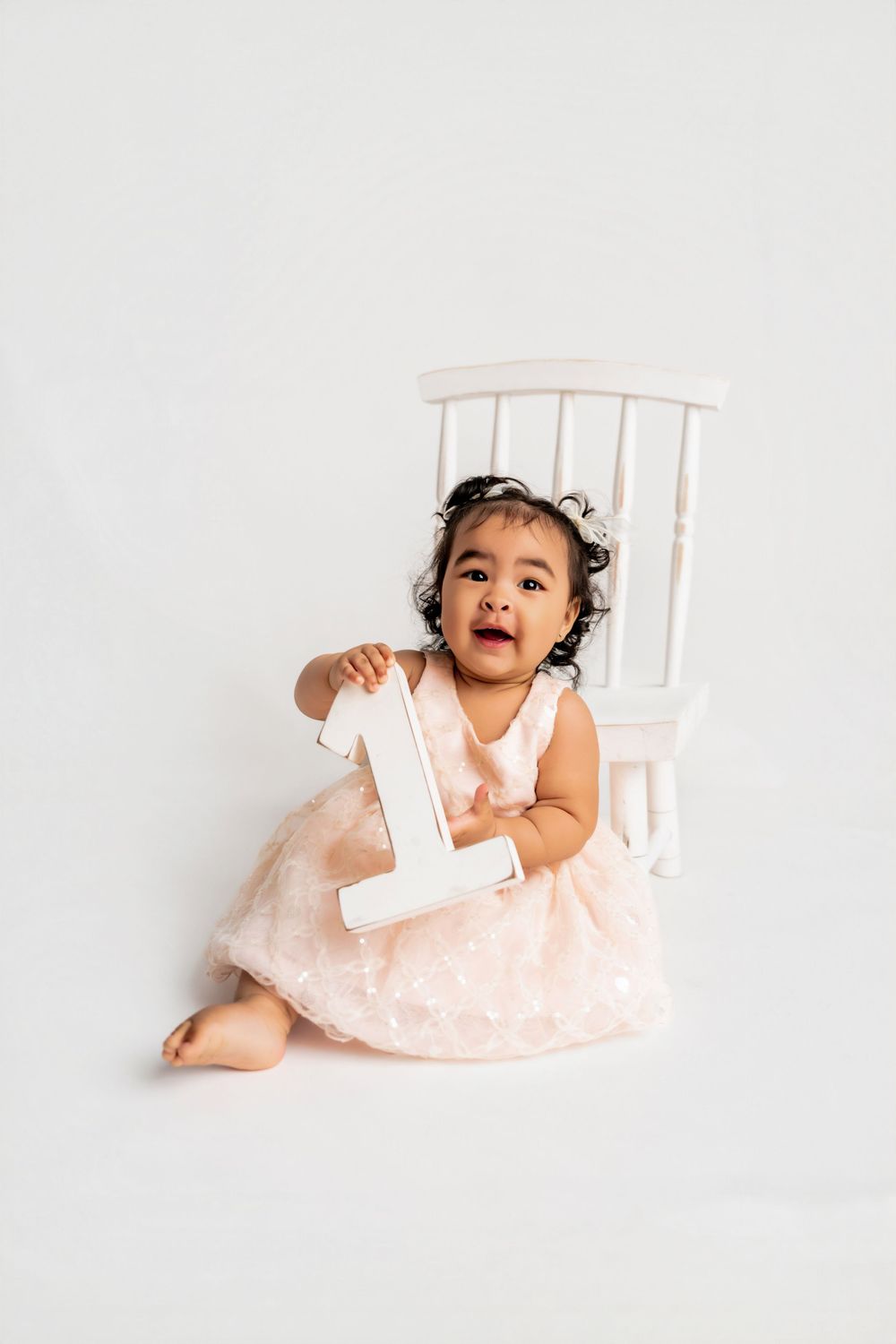 Cute baby girl in pink dress holding number 1, sitting by white chair, celebrating first birthday.