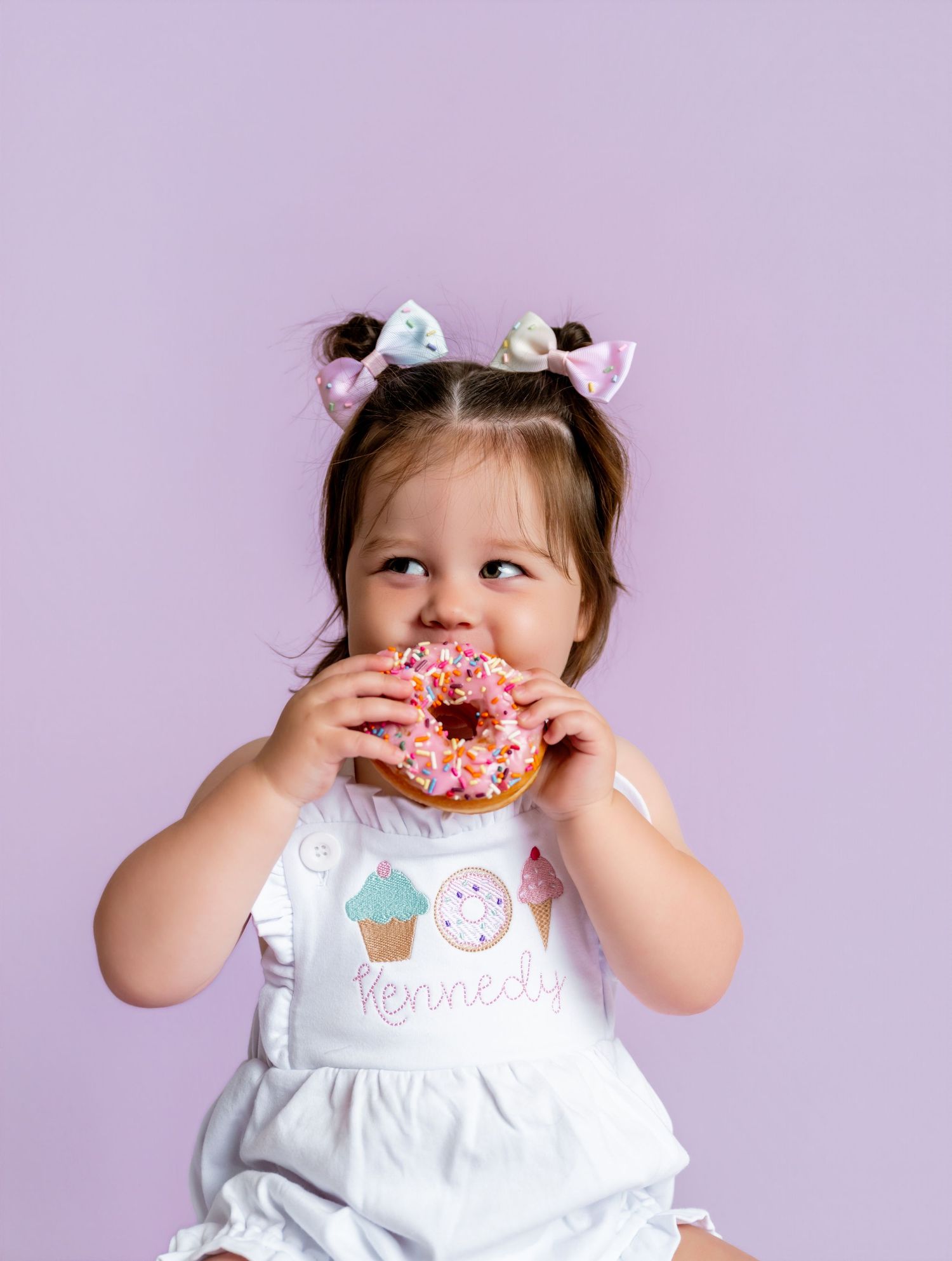 Toddler girl in white romper with pigtail bows eating a pink sprinkle donut against lavender background.