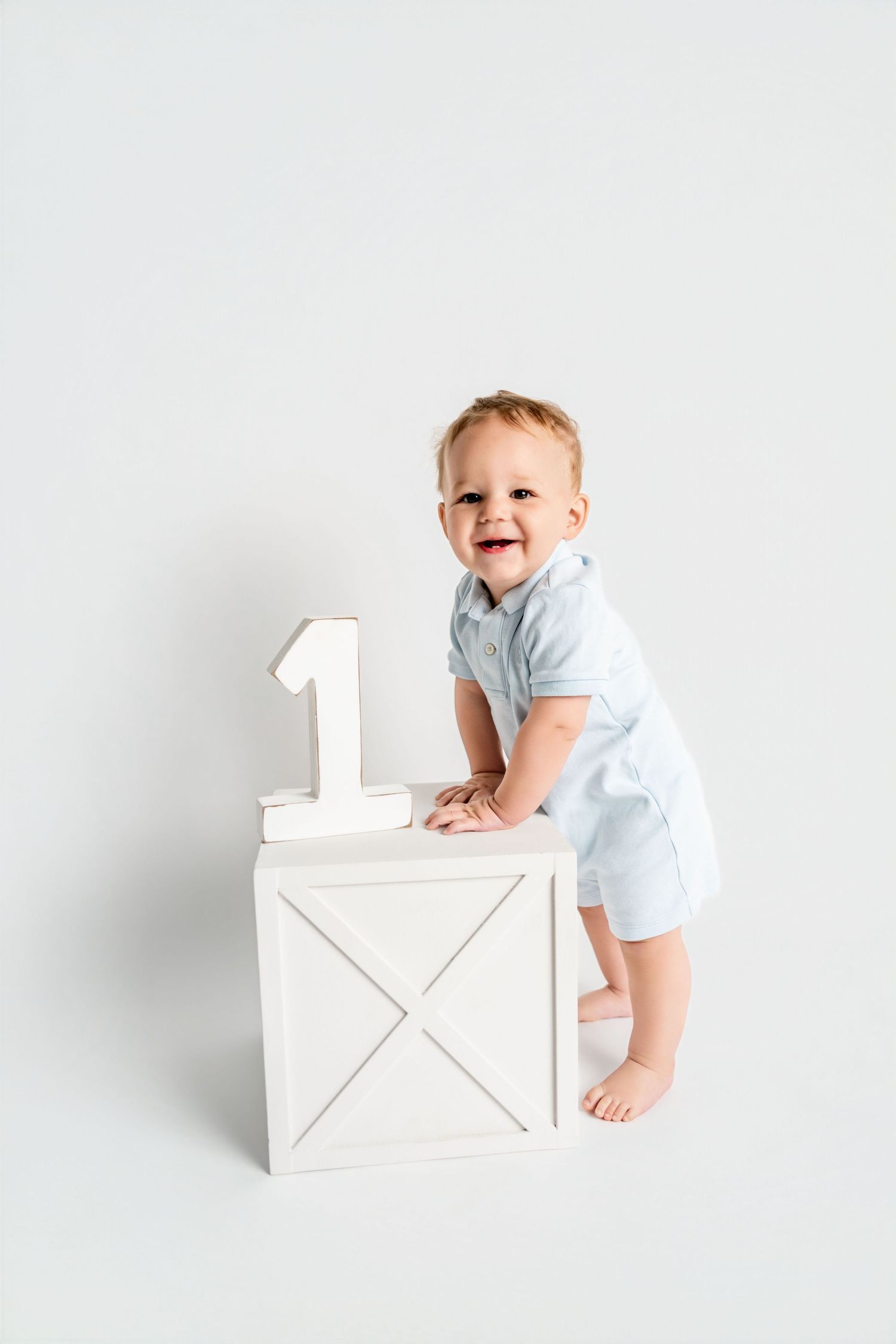 Smiling baby boy in light blue outfit standing by white wooden box with number 1 for first birthday photo.