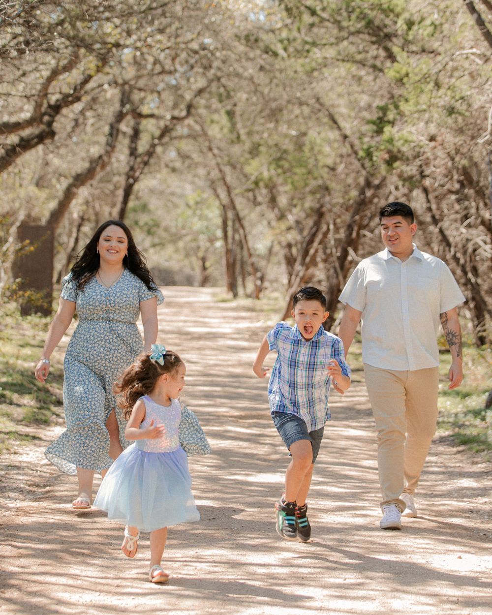 Family plays and runs together on a tree-lined dirt path in coordinated blue and white outfits.