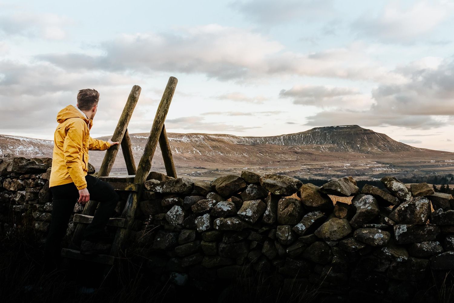 A person in a yellow jacket stands on dark volcanic rocks against a cloudy sky at sunset.