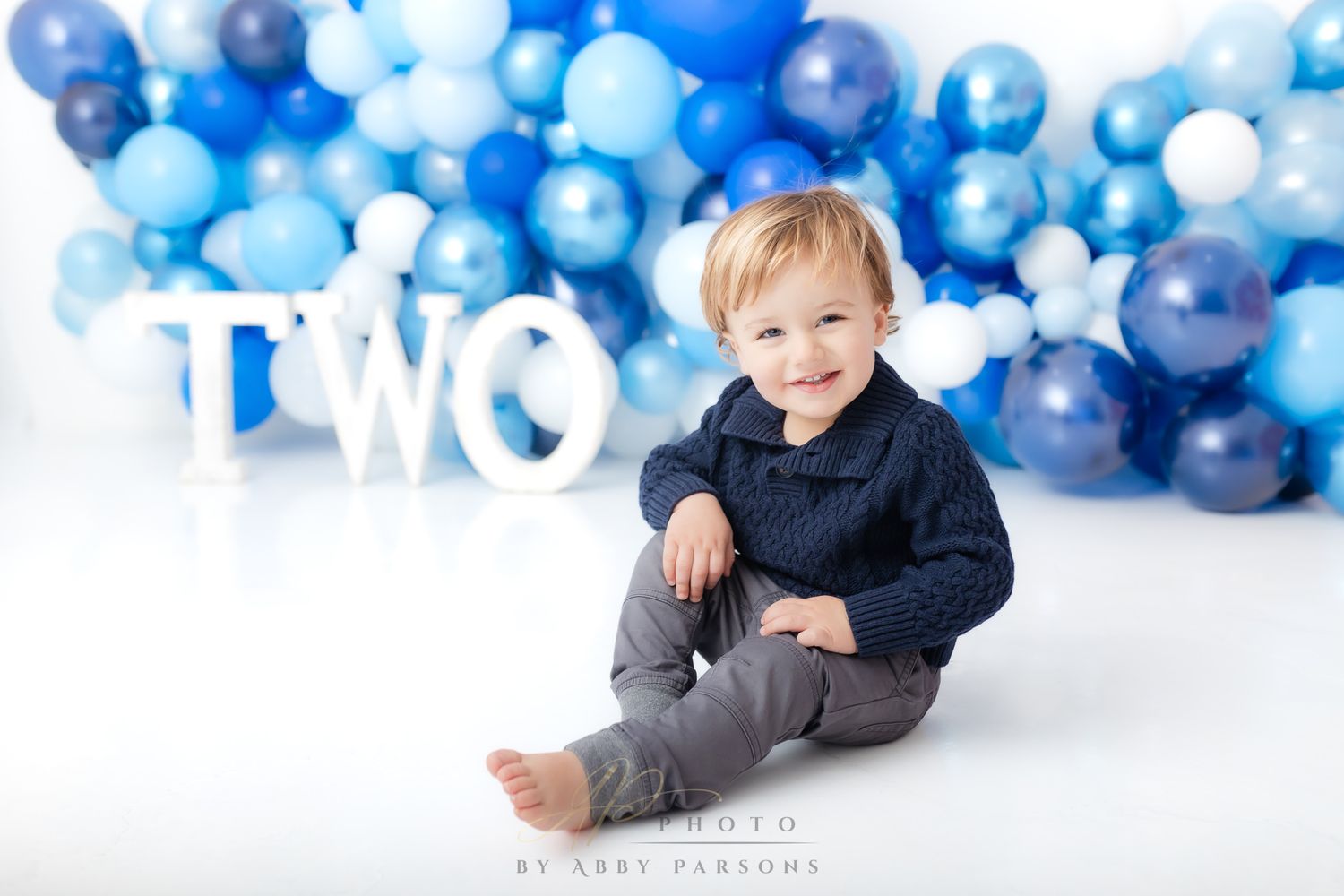 Small child sits barefoot on white floor with blue and white balloon arch backdrop and white TWO letters behind.