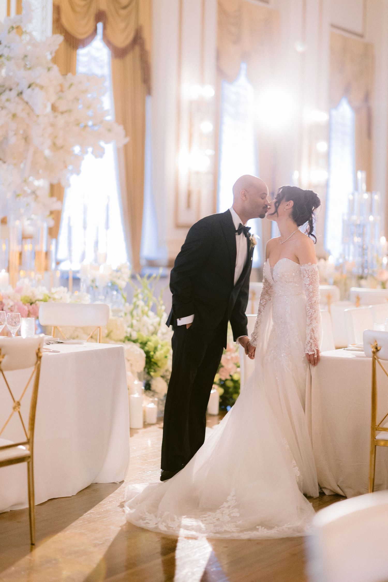 Romantic wedding couple shares intimate moments in an elegant white ballroom with gold chairs and floral decorations.