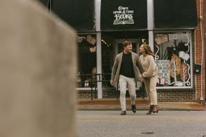A couple walks down the sidewalk holding hands in front of a storefront on a sunny day.