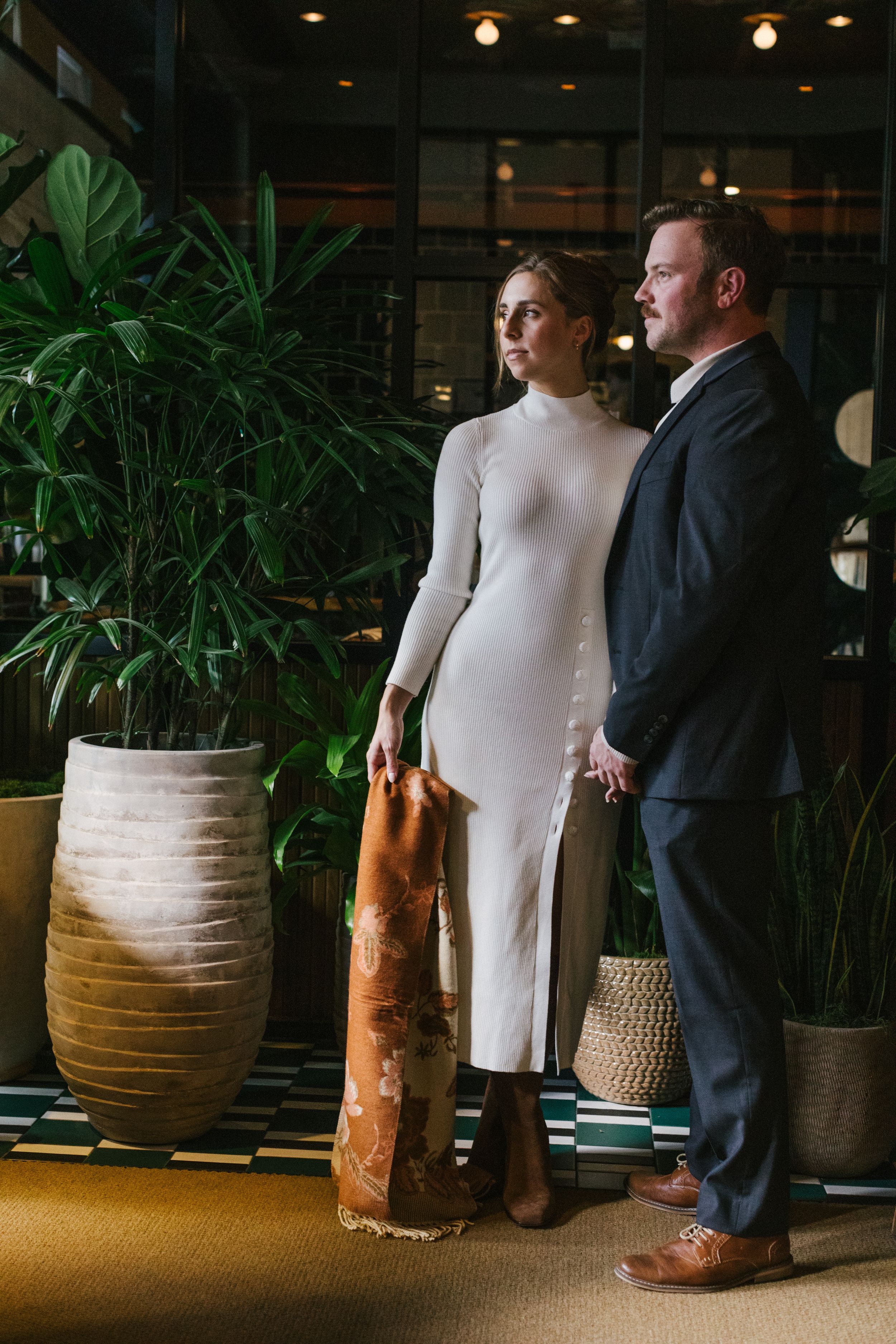 A couple stands in an elegant hotel lobby with modern decor featuring tall plants and decorative vases.