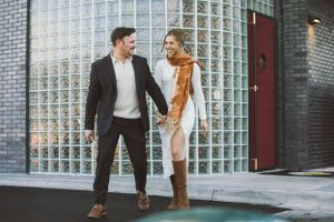 Stylishly dressed couple stands together holding hands near a glass block wall and red door.