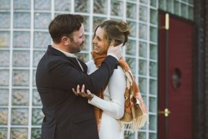A romantic couple embraces outside a building with glass block windows and a red door on an autumn day.