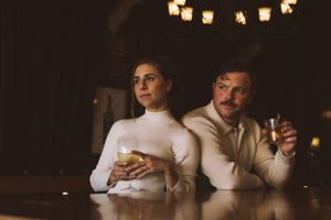 A retro-styled couple in white attire sits at a dimly lit bar holding cocktail glasses with warm lighting above.