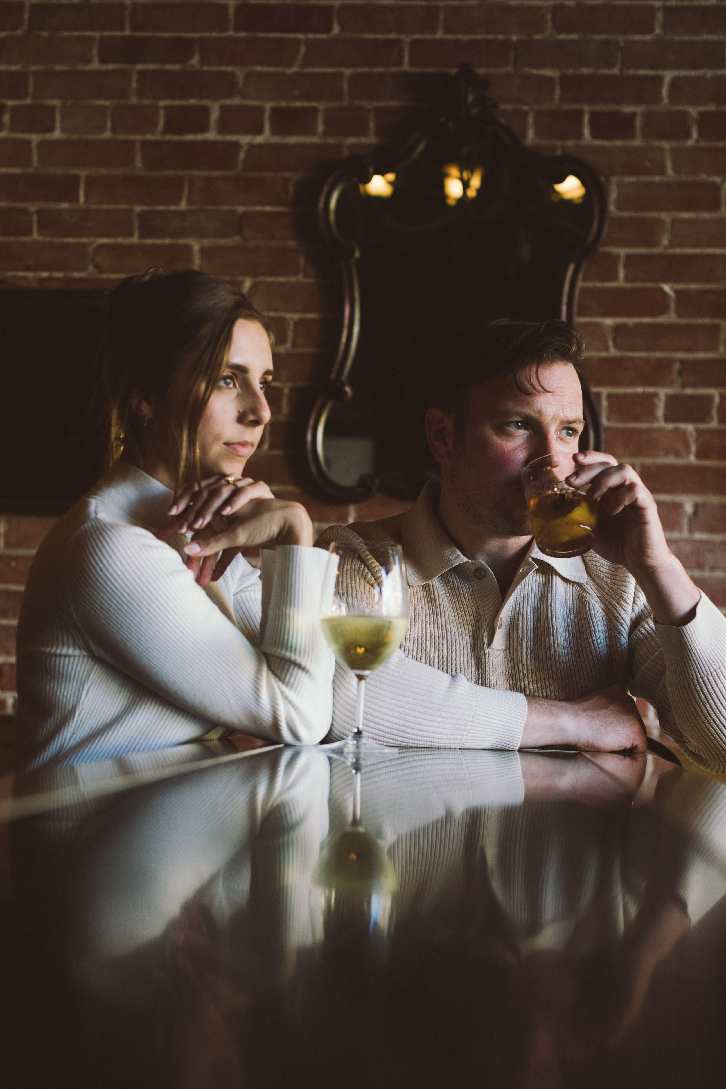Two people in white sweaters sit at a reflective table with cocktails in a dimly lit room with exposed brick walls.