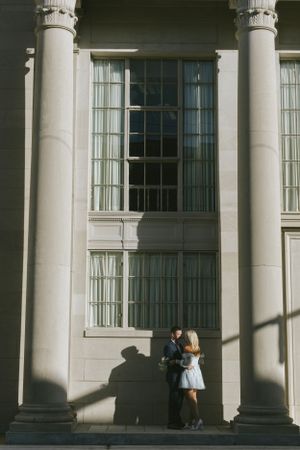 A couple stands between tall white columns of a classical building, casting long shadows in the sunlight.