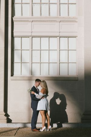 Romantic couple embraces against white wall with a heart-shaped shadow cast beside them in vintage-style photograph.