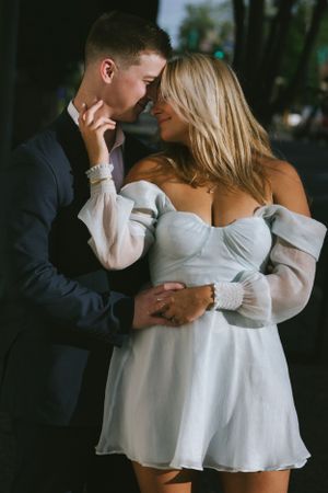 A romantic couple embraces in formal attire, with the woman wearing a flowy white off-shoulder dress and man in dark suit.
