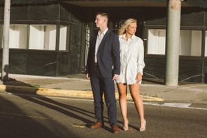 A couple in formal attire poses together in an urban setting with concrete pillars and sunlit background.