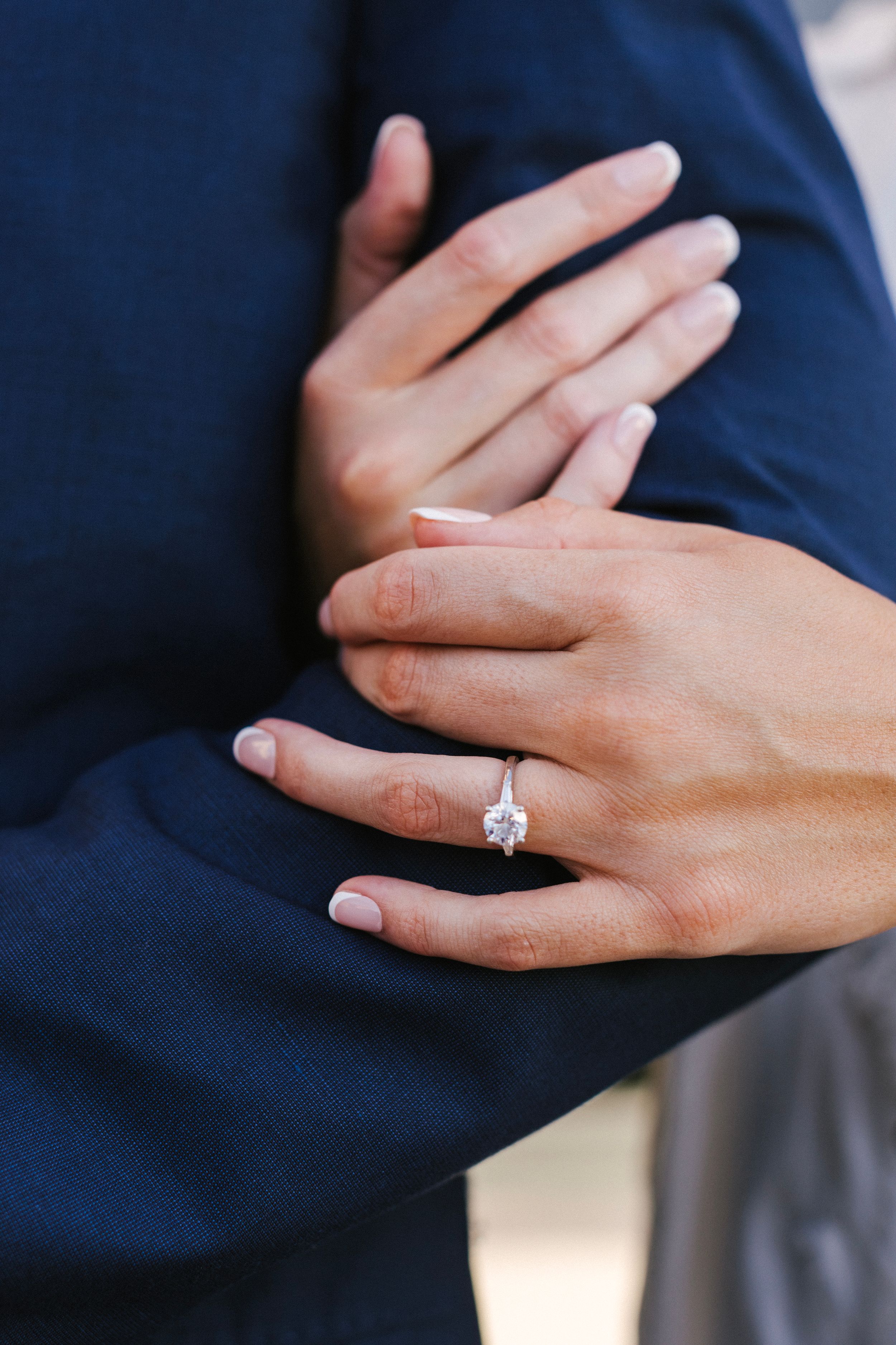 A close-up of hands showing off a sparkling engagement ring on a finger against a navy blue suit jacket.