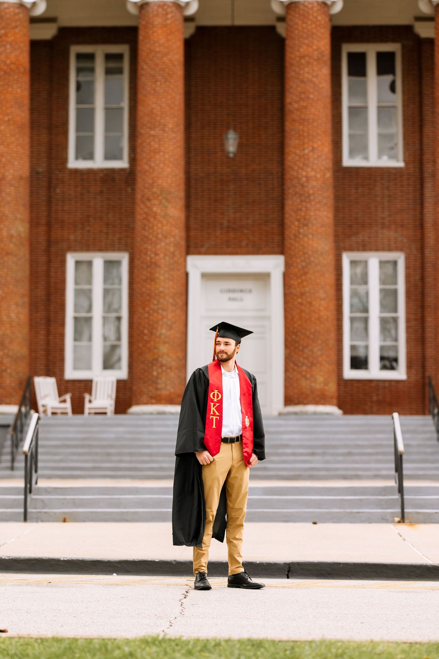A graduate in cap and gown stands in front of a brick building with white columns.