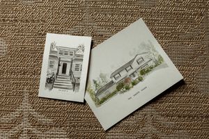 Two hand-drawn architectural sketches of homes on cards, one black-and-white and one color watercolor, displayed on textured fabric.
