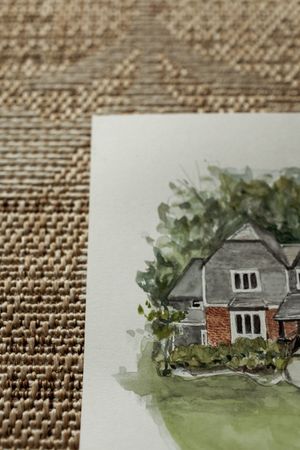Watercolor painting of a charming brick and gray house surrounded by lush green trees, placed on a woven texture surface.