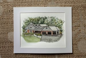 Watercolor painting of a large suburban house with garage, lush green trees, matted in white frame.