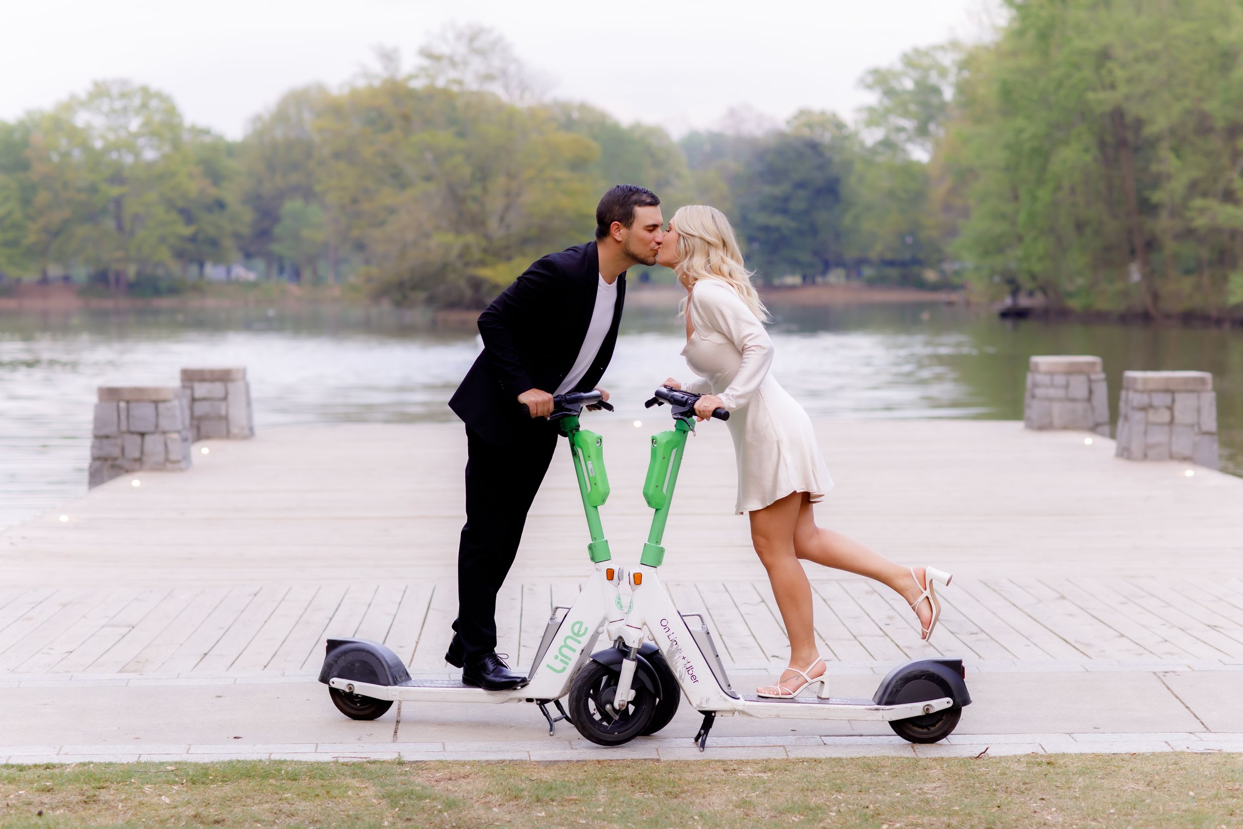 Playful couple poses on electric scooters in casual wedding attire by waterfront.