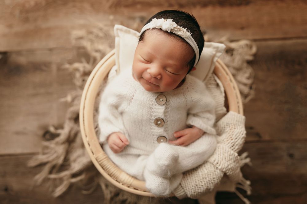 A sleeping newborn wearing a white knit outfit rests peacefully in a round wooden basket with cozy textiles.