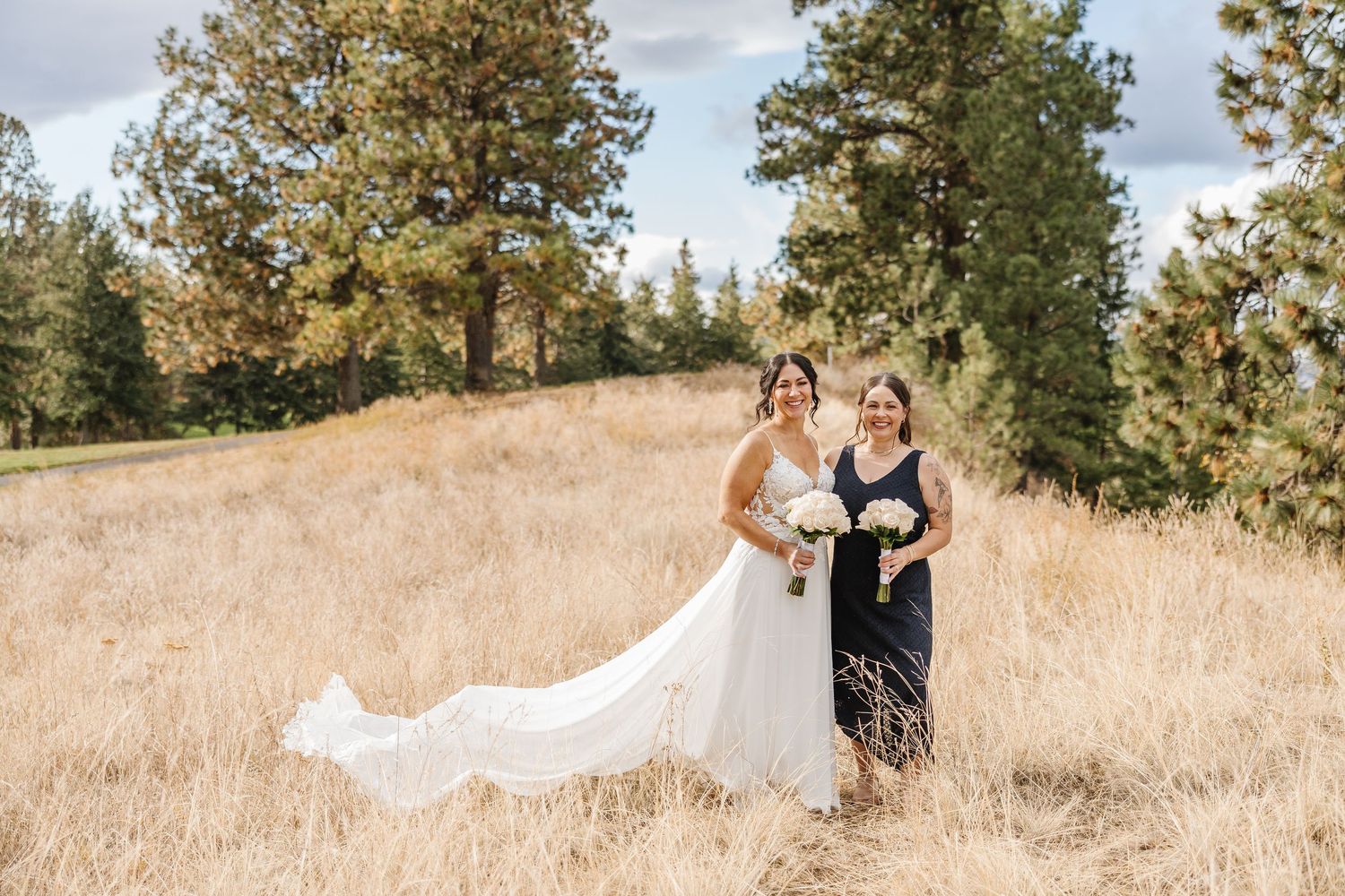 A couple in wedding attire walks through a golden field with tall grass and trees on a sunny day.