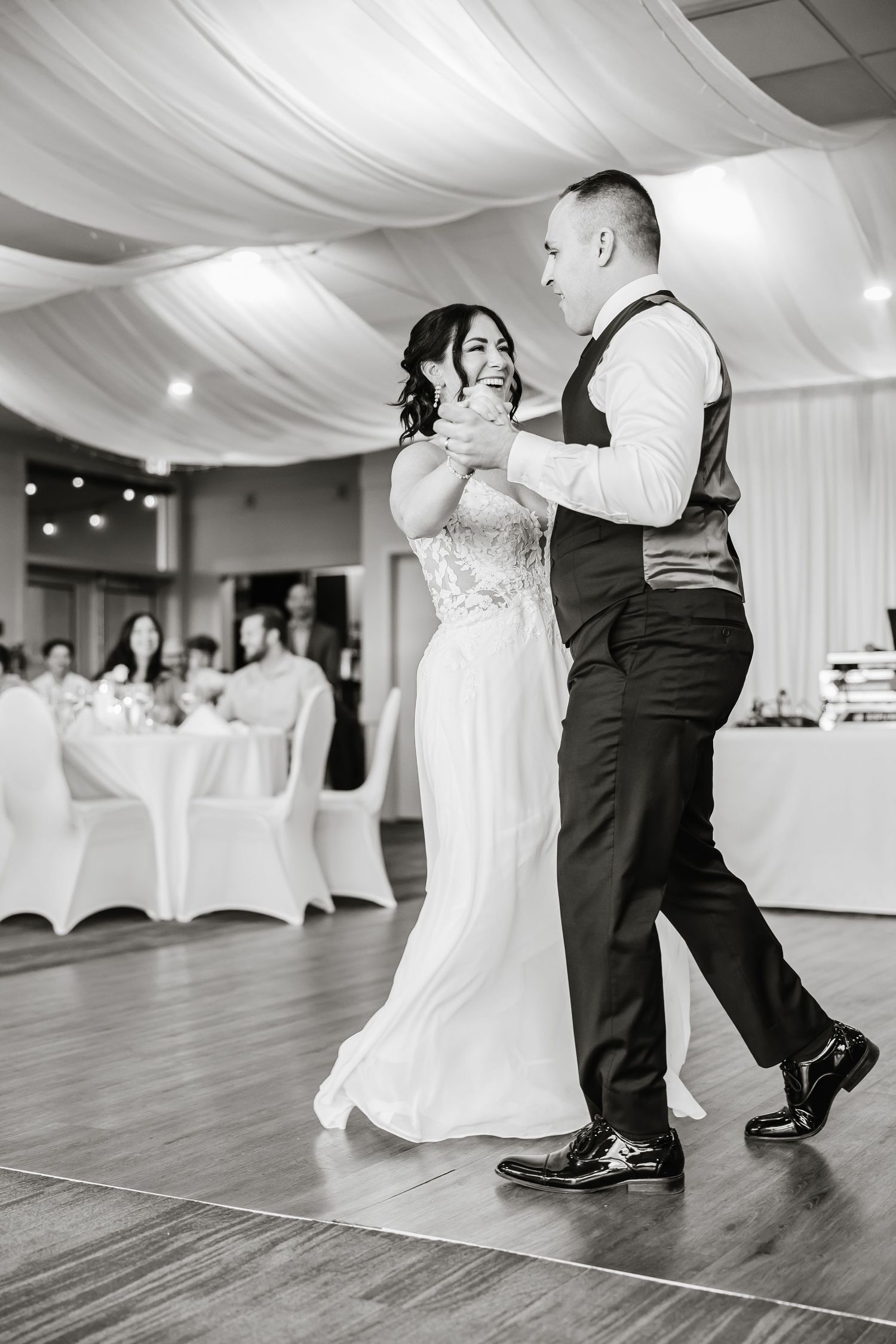 Wedding guests dance together on a wooden floor under elegant white draped ceiling decor.