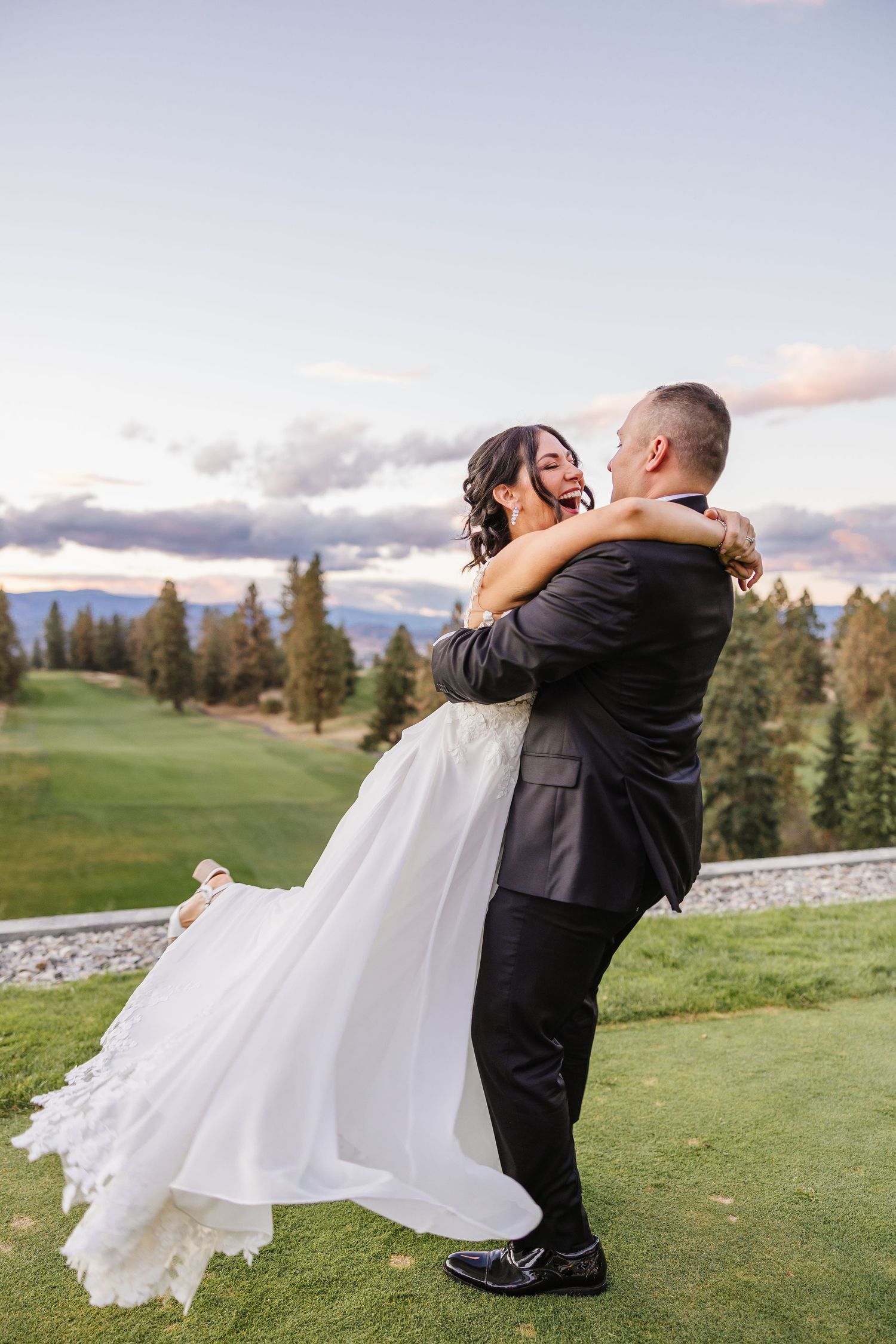 A romantic wedding couple embraces on a scenic golf course with evergreen trees and cloudy skies in the background.