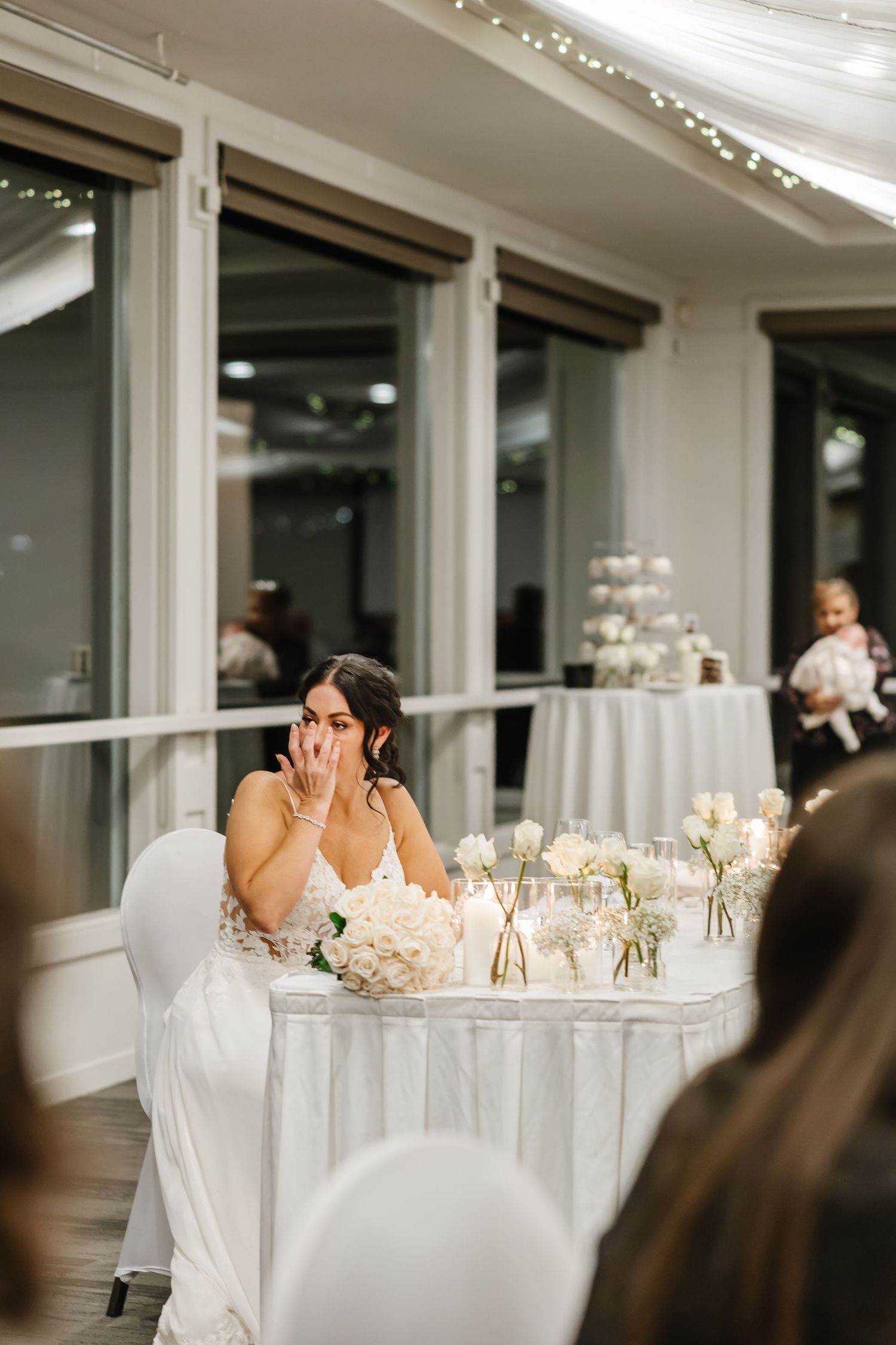 A bride in a white dress sits at an elegantly decorated reception table with floral centerpieces and string lights overhead.