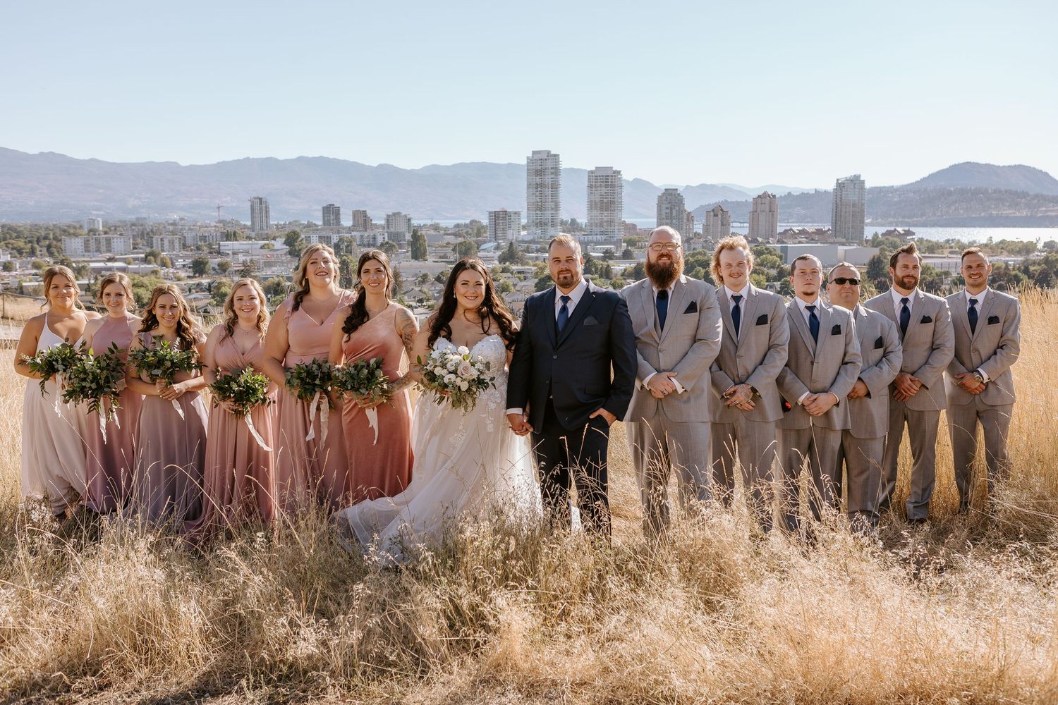 Wedding party stands together on a hillside overlooking a scenic mountain and city landscape in the background.