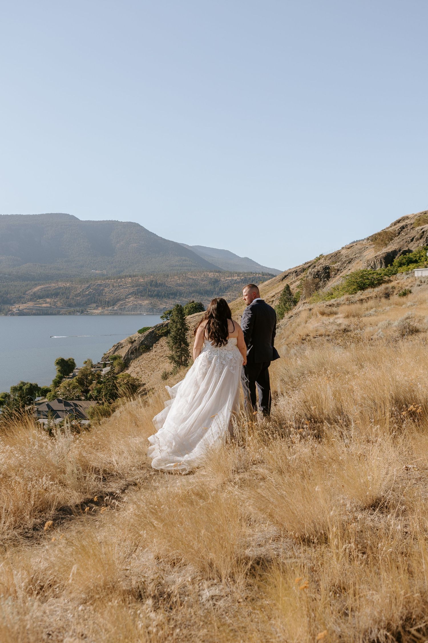 Newlyweds walk along scenic coastal hillside with mountain and ocean views in background.