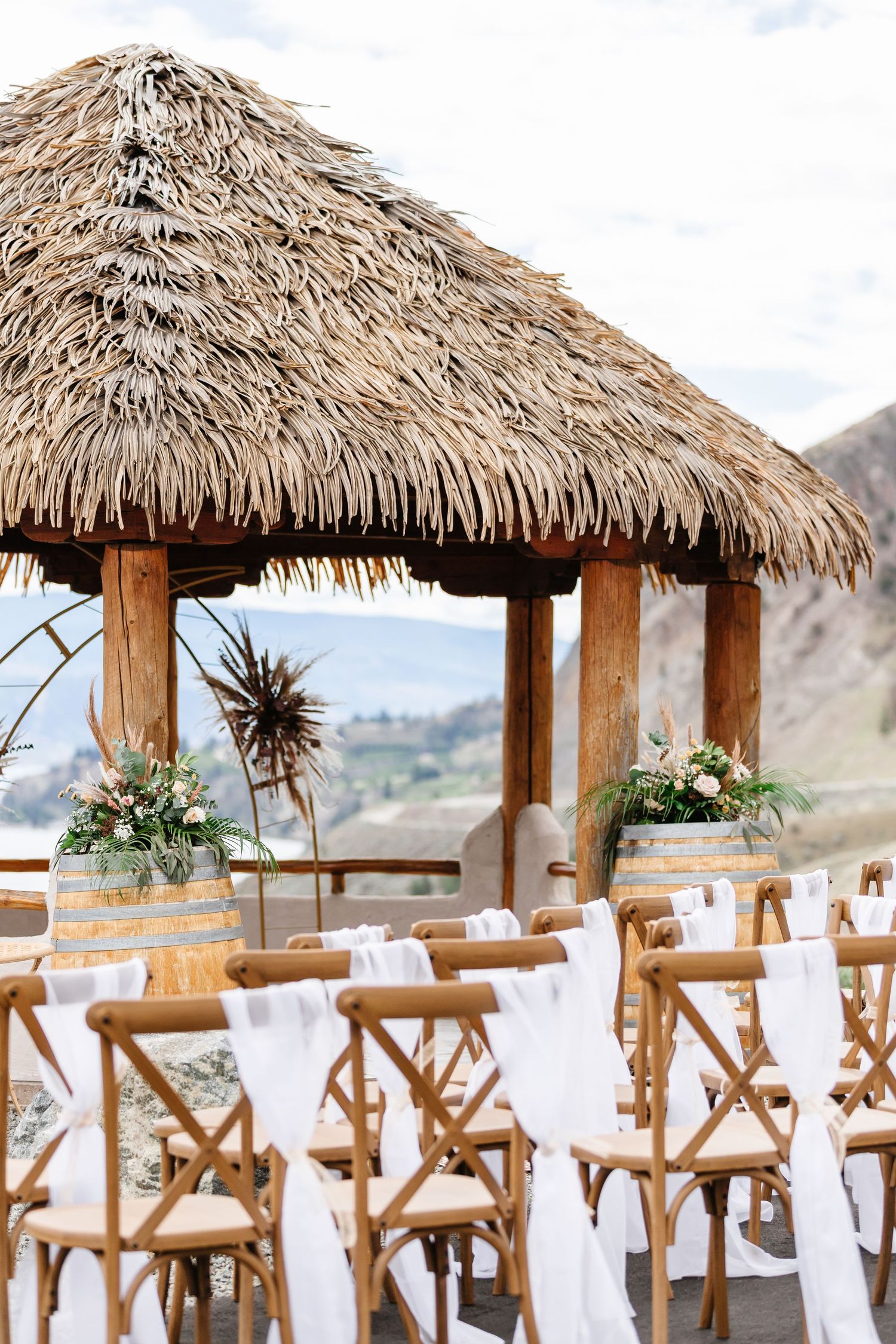 Thatched-roof pavilions with wooden chairs set up for an outdoor wedding ceremony with mountain views.