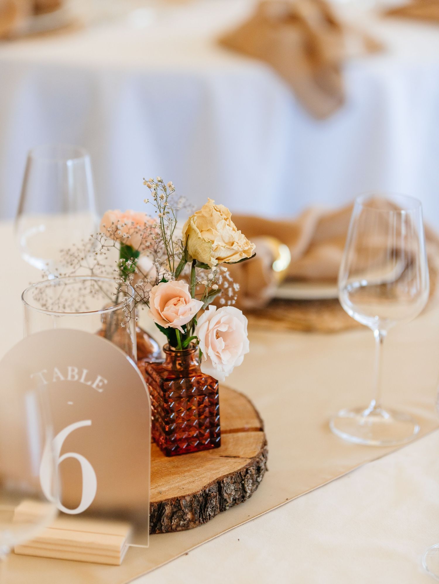 Rustic wedding table centerpiece with peach roses in amber vase on wood slice with table number six and wine glasses.