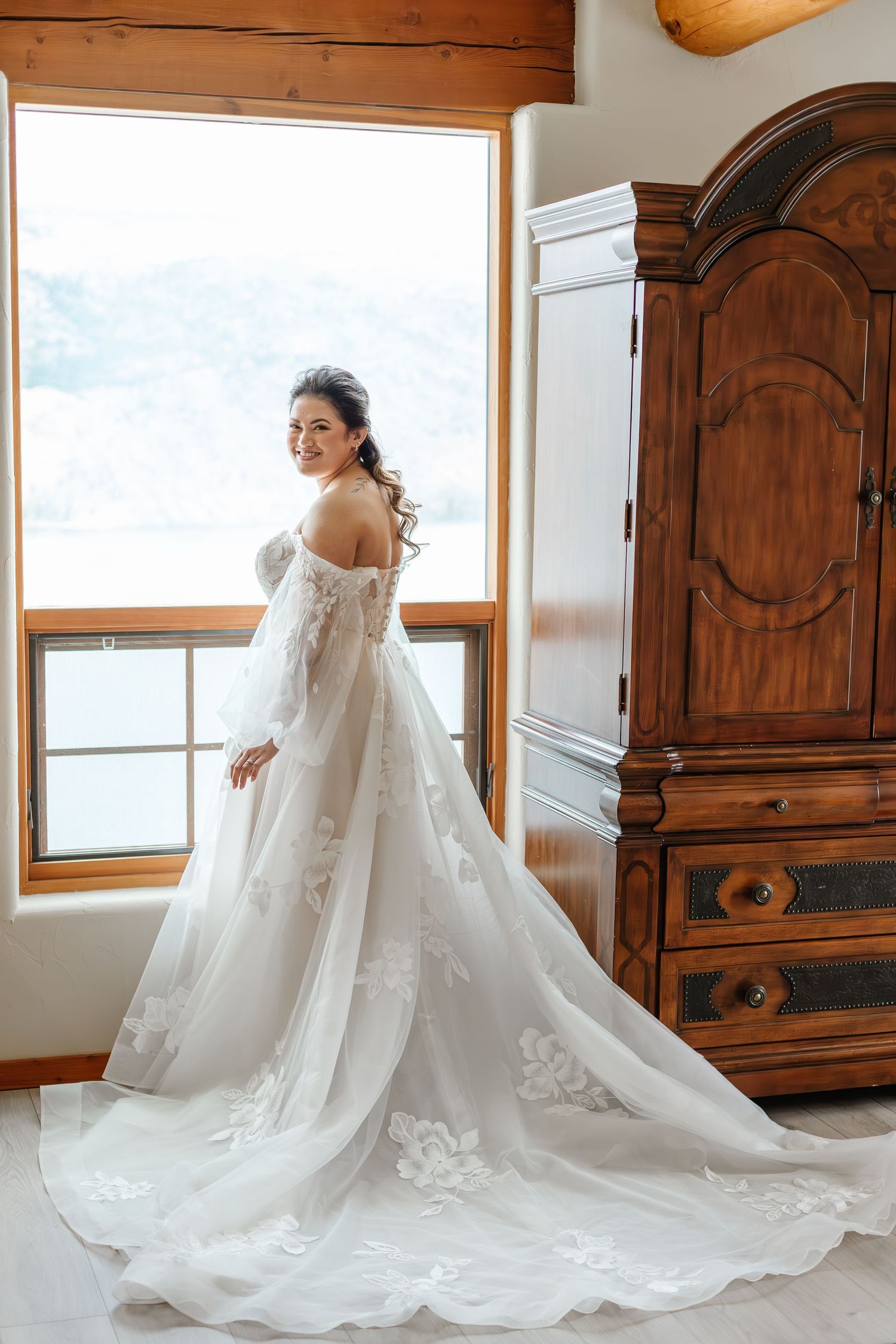A bride in a flowing white wedding gown with a dramatic cathedral-length lace train stands in front of wooden furniture.