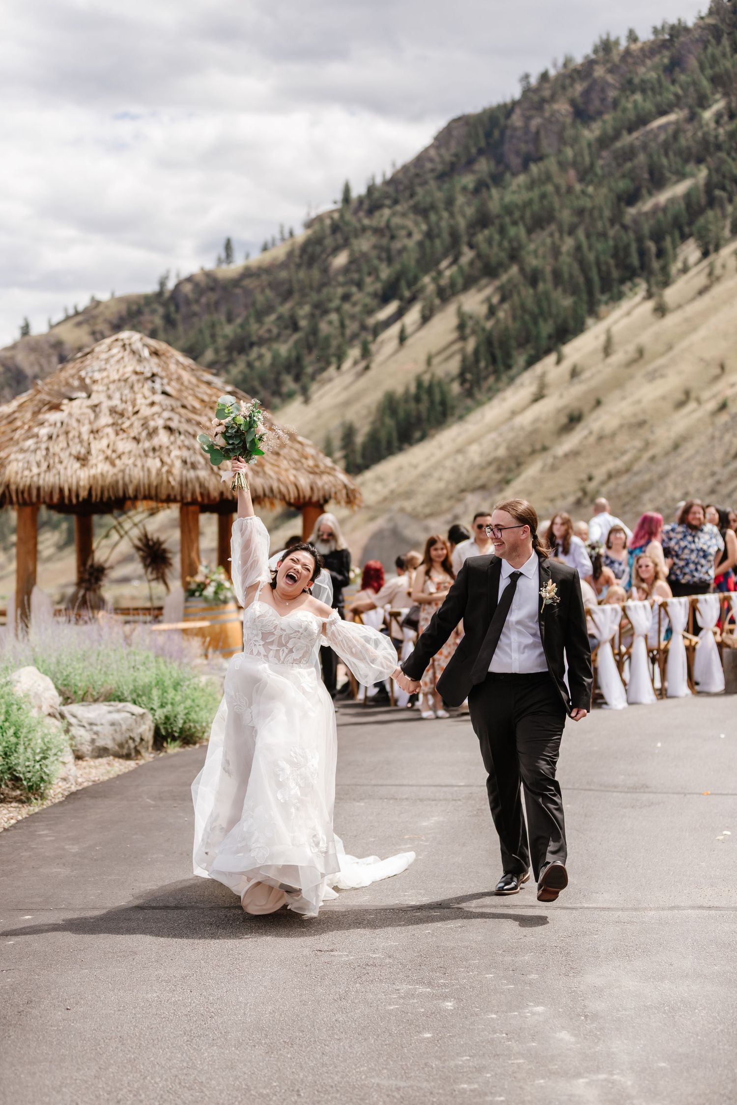 Newlyweds celebrate with joyful dance moves on a mountain path with a thatched-roof gazebo in the background.
