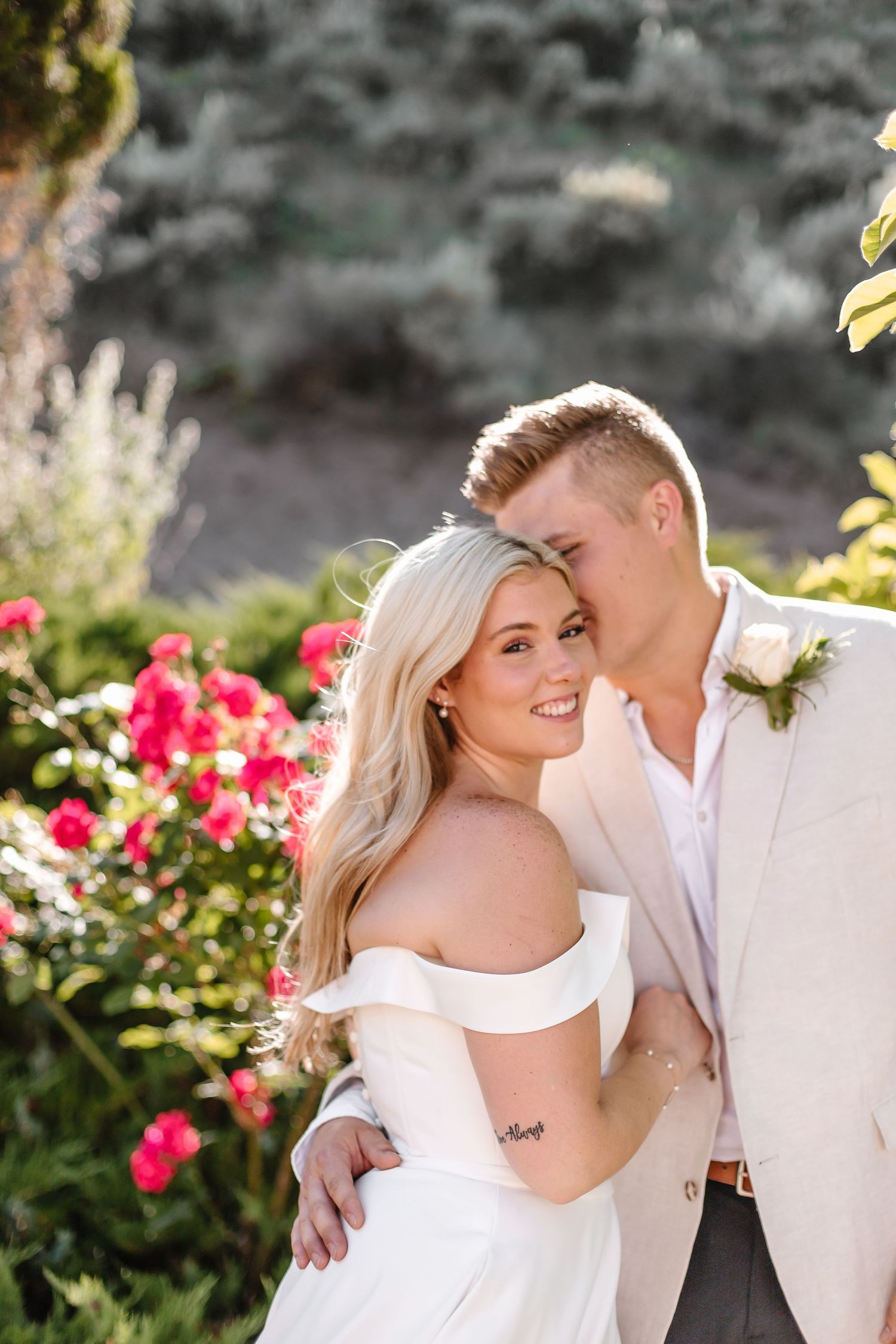 Intimate wedding portrait with couple embracing among blooming pink roses in a garden.