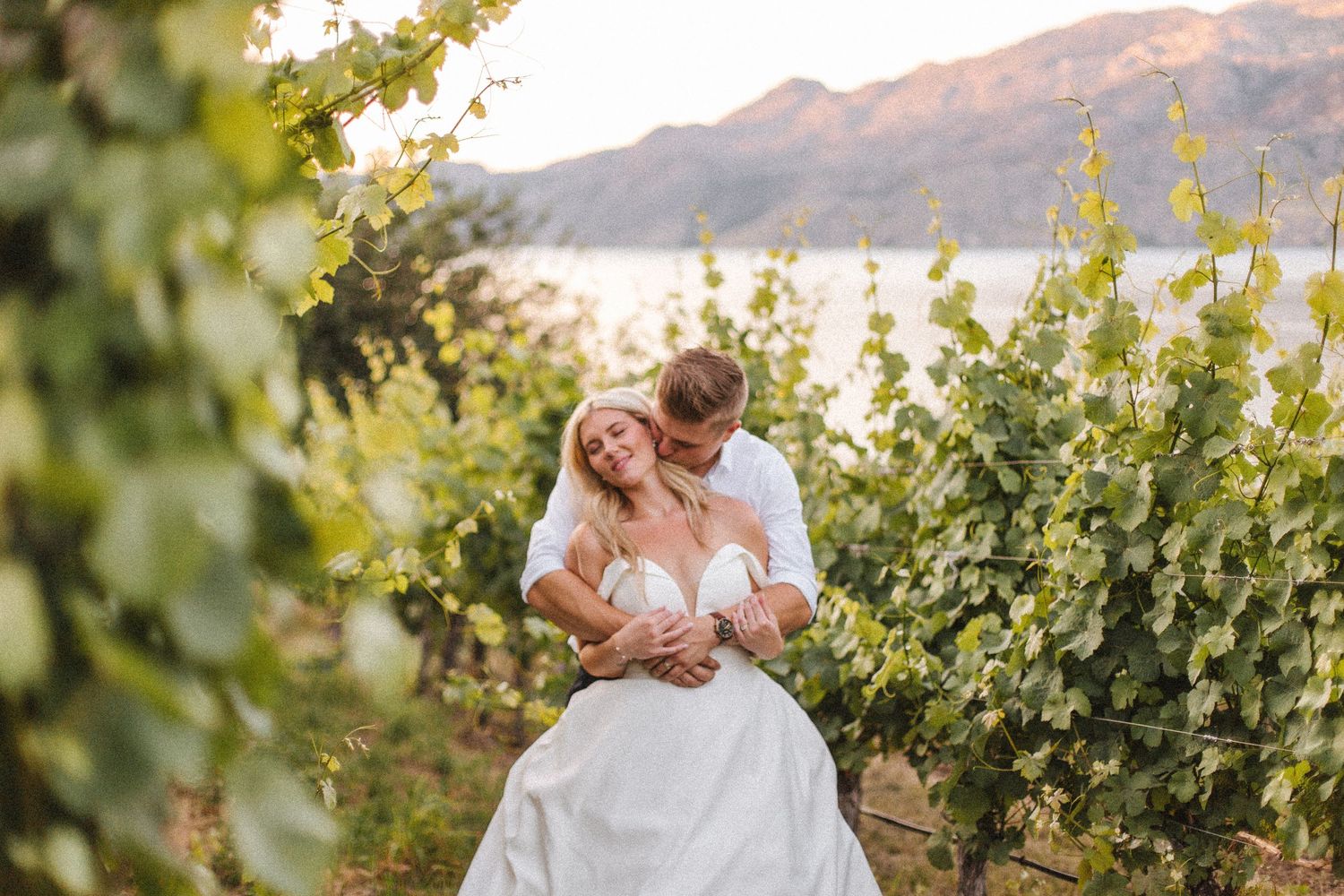 A couple embraces in a sunlit vineyard with mountains visible in the background.