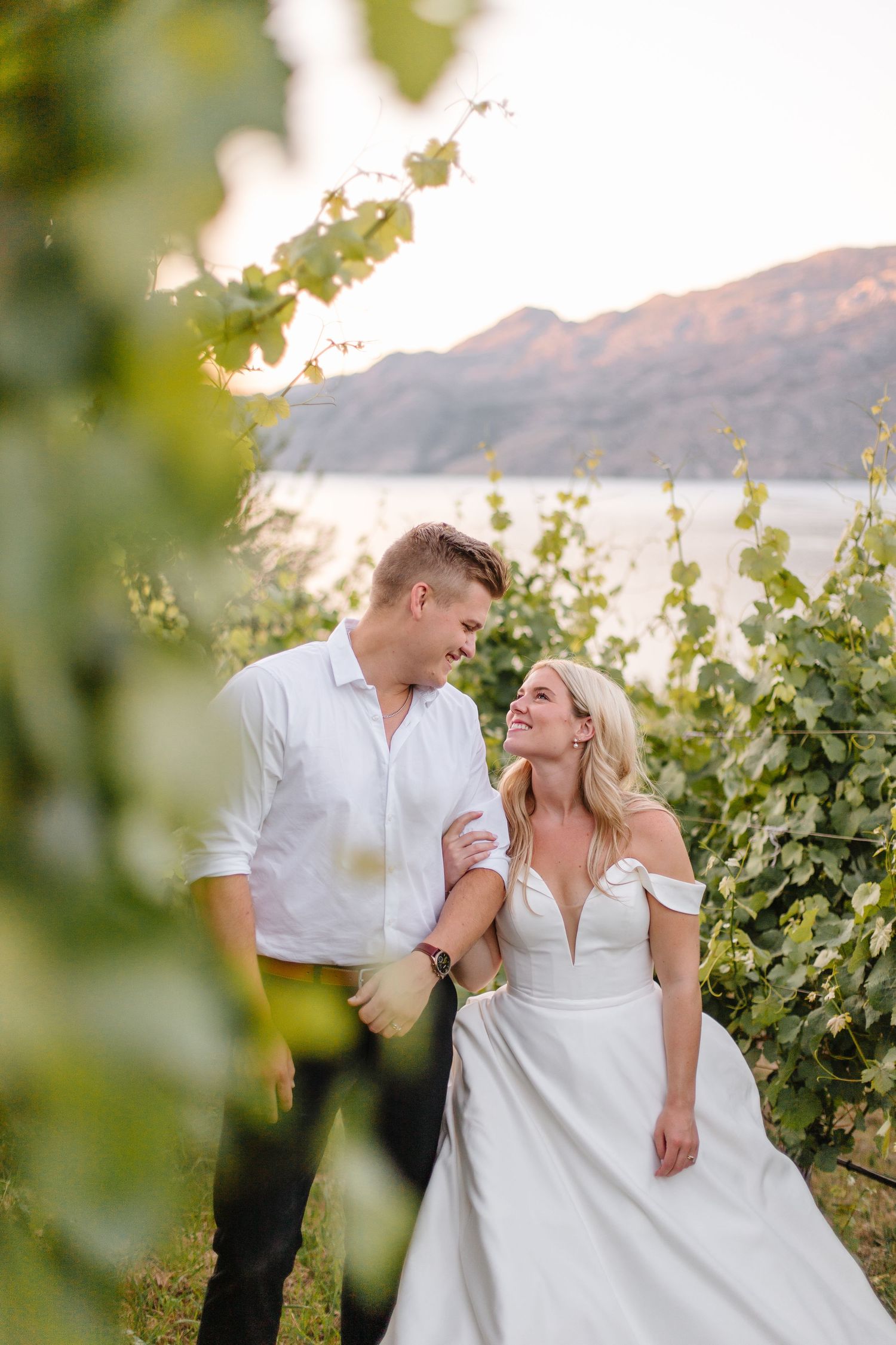 A couple shares a romantic moment among grapevines overlooking a scenic lake at sunset.