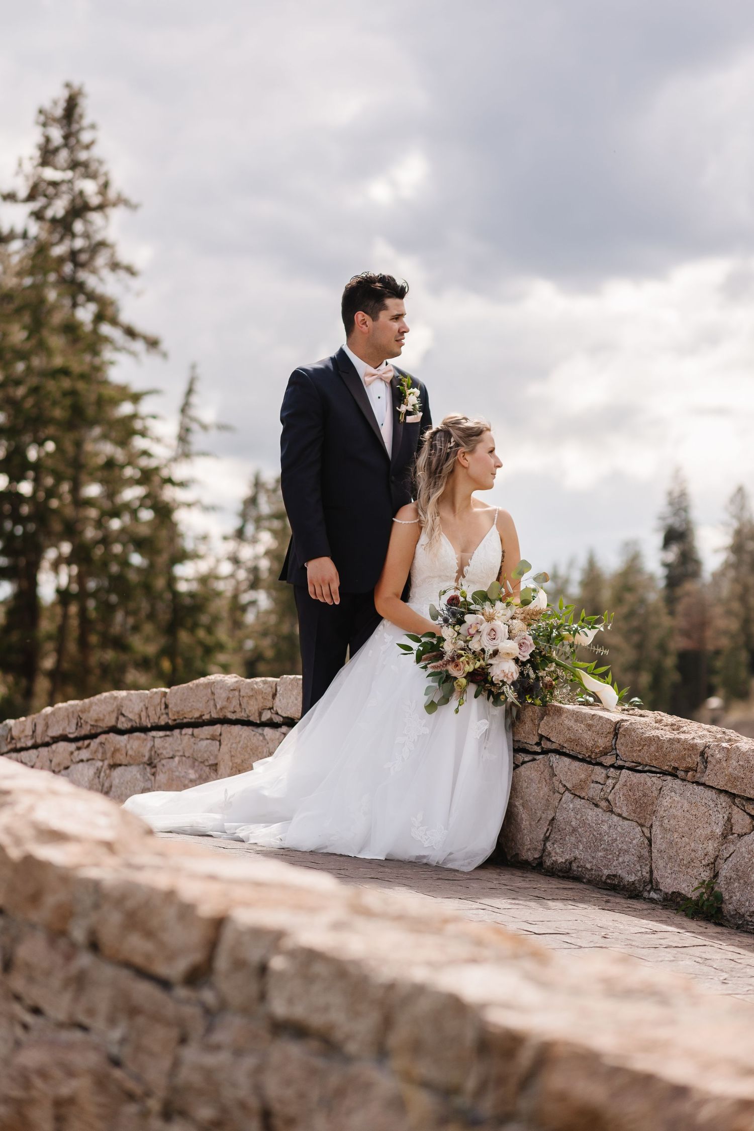 A couple in wedding attire sits together on a rustic stone wall with evergreen trees and cloudy skies in the background.