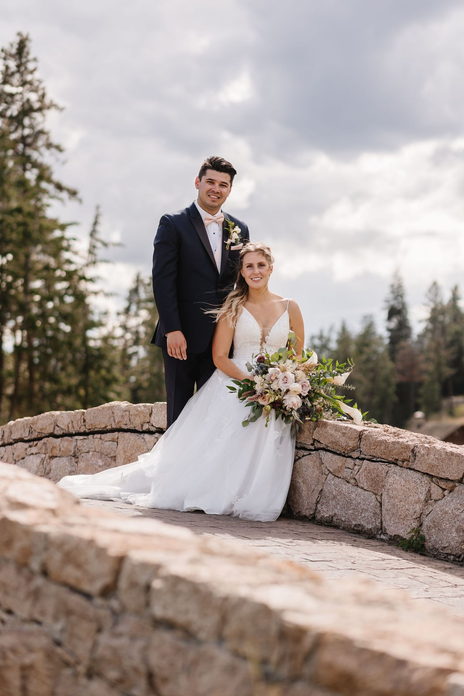 A couple in wedding attire pose together on a stone wall with evergreen trees and cloudy skies in the background.
