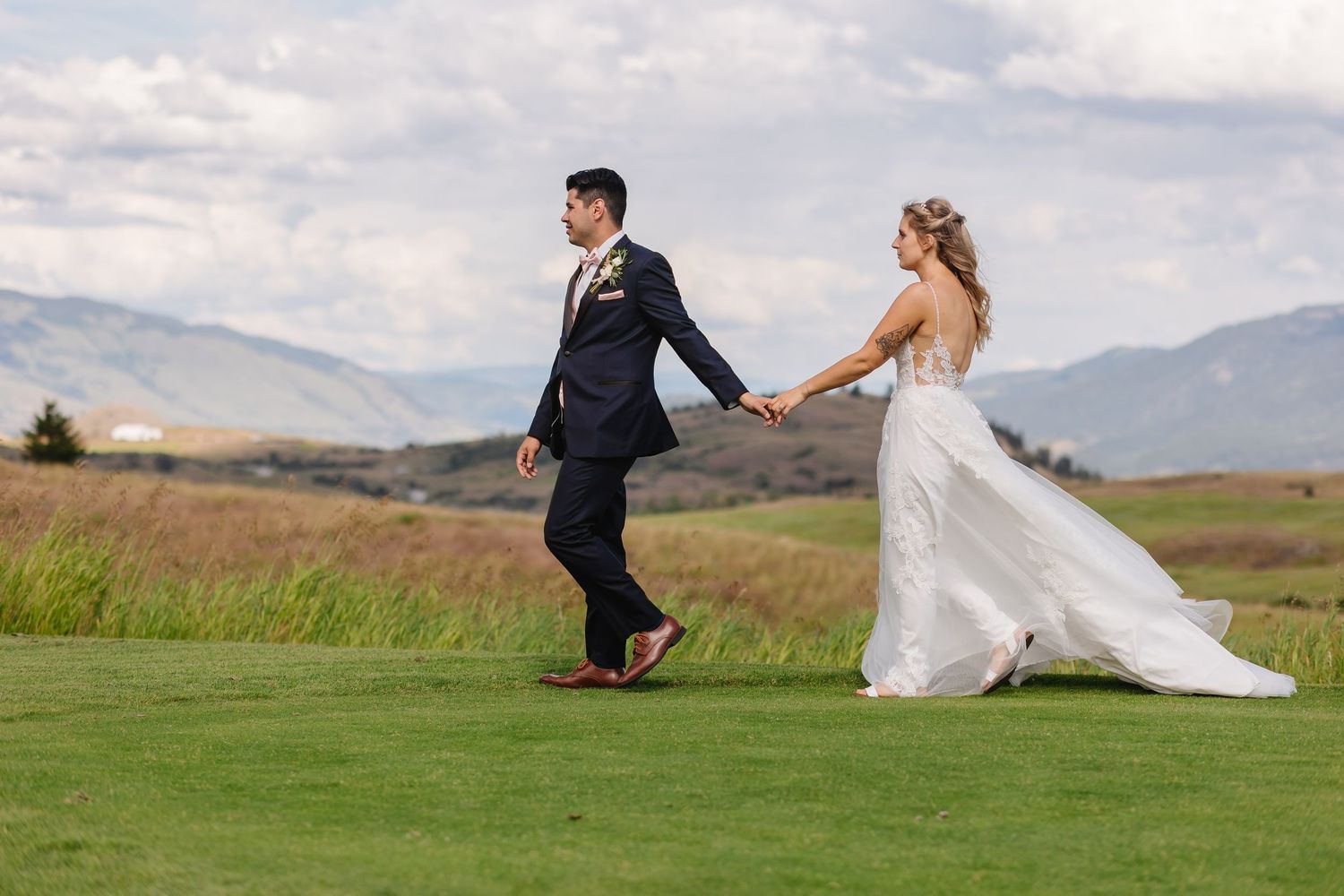 A couple in wedding attire walks hand in hand across a scenic golf course with mountains in the background.