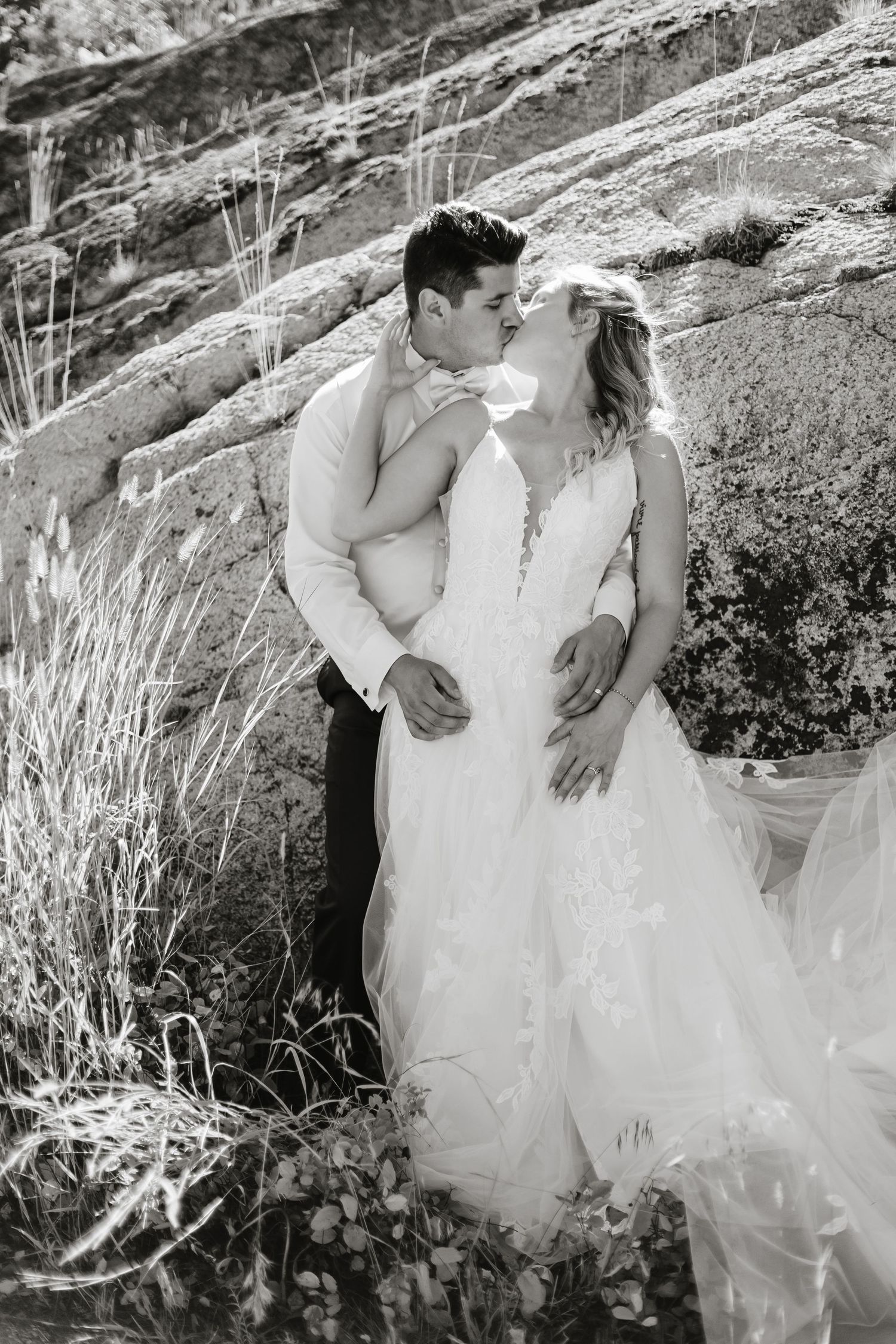 A romantic black and white wedding photo of a couple embracing outdoors against a rocky hillside with flowing grasses.