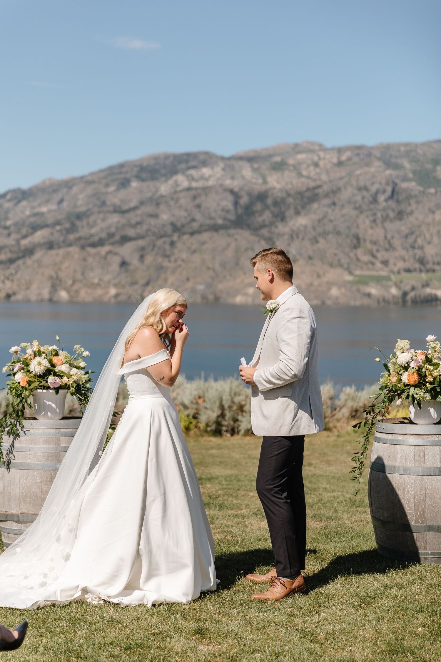Wedding ceremony setup with wine barrels and floral arrangements against a mountainous lakeside backdrop.